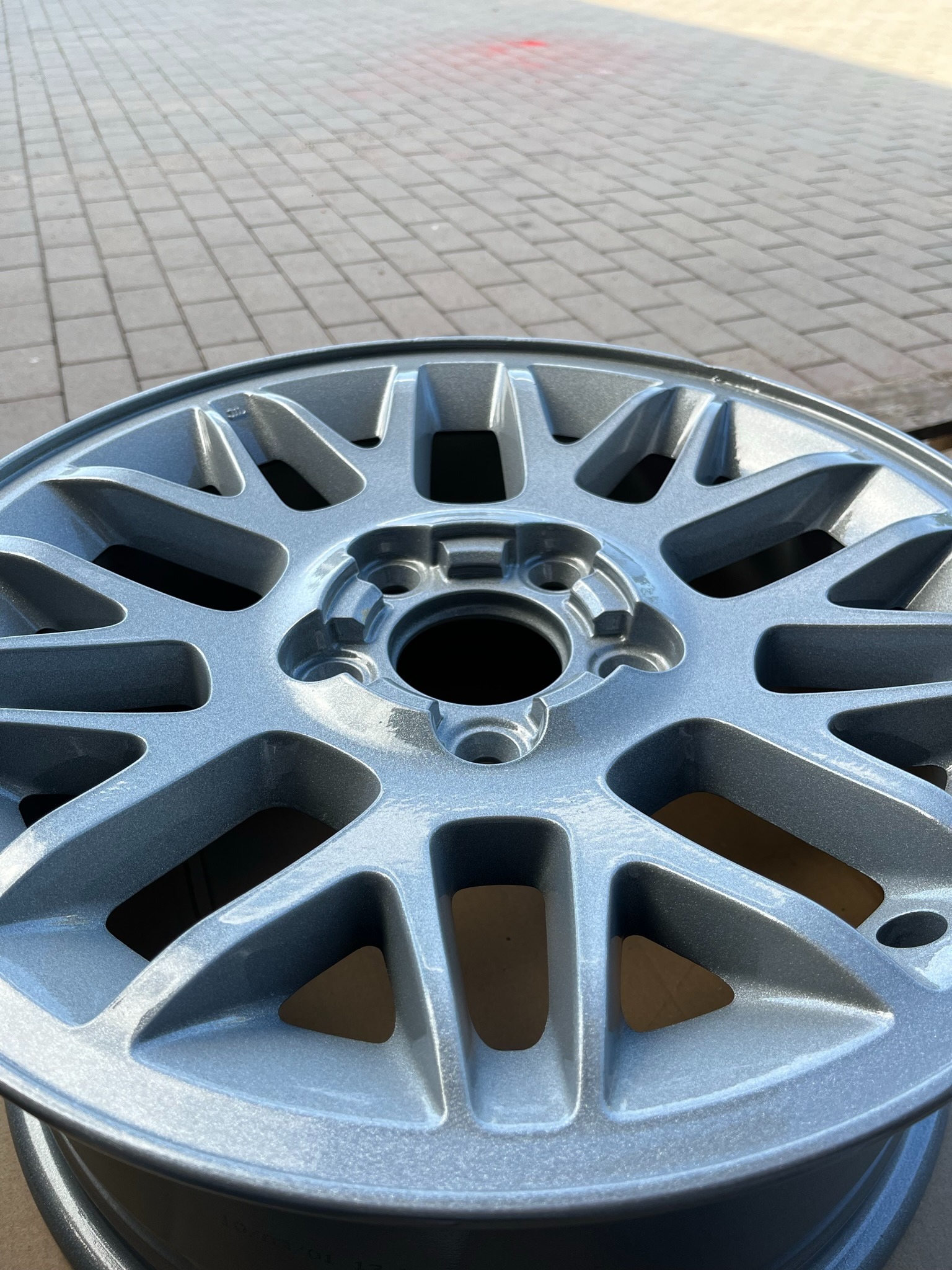 Błyszcząca, srebrna felga aluminiowa po malowaniu proszkowym, widoczna na tle kostki brukowej.