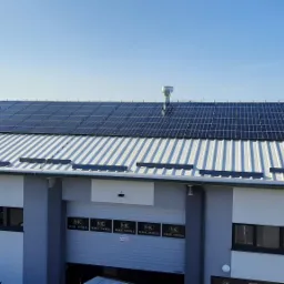 Instalacja paneli fotowoltaicznych na dachu hali przemysłowej z blachy trapezowej, widoczne kominy wentylacyjne i logo firmy na bramie wjazdowej.