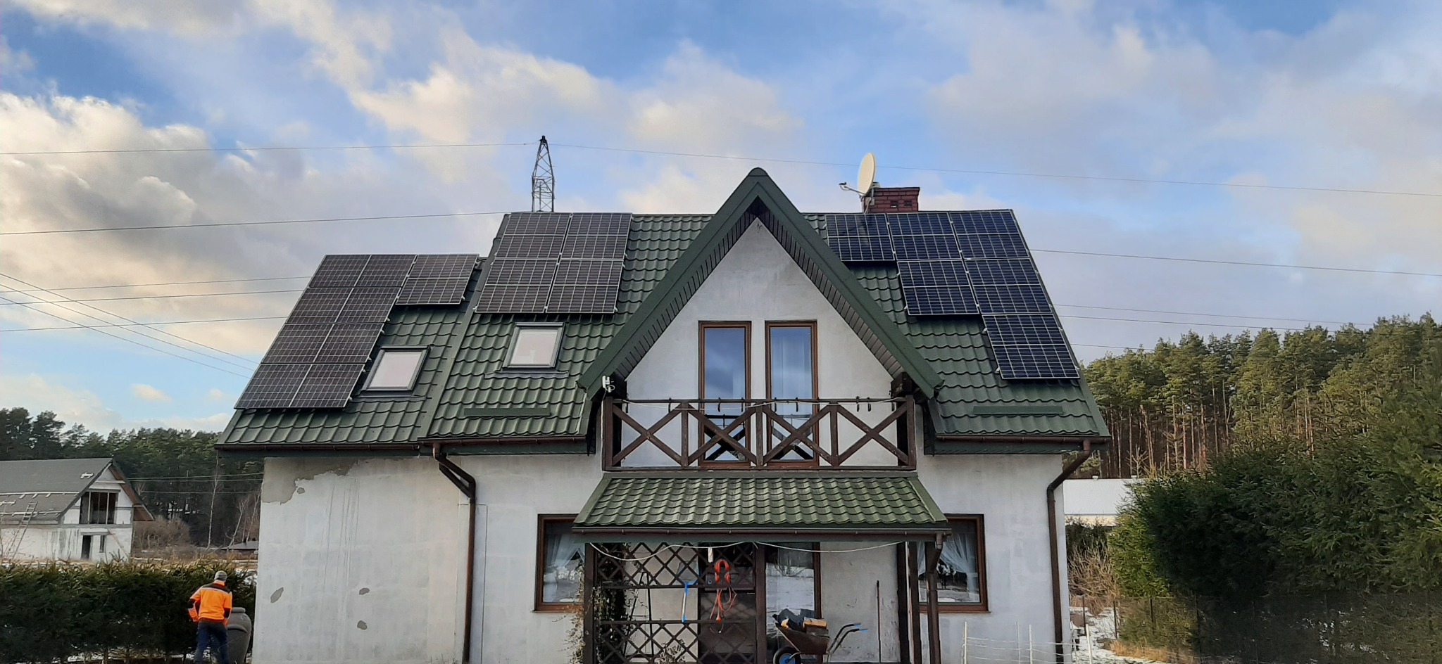 Dom jednorodzinny z zainstalowanymi panelami słonecznymi na dachu pokrytym zieloną dachówką, widoczne okna dachowe i balkon z drewnianą balustradą, na pierwszym planie osoba w pomarańczowej kurtce.