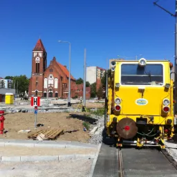 Żółta maszyna torowa z widocznym operatorem w kamizelce odblaskowej na tle placu budowy z czerwonym hydrantem i kościołem w oddali.