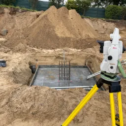 Geodezyjny niwelator Leica na żółtym statywie, ustawiony obok wykopu z betonową płytą fundamentową i zbrojeniem, w tle hałda ziemi i ogrodzenie.