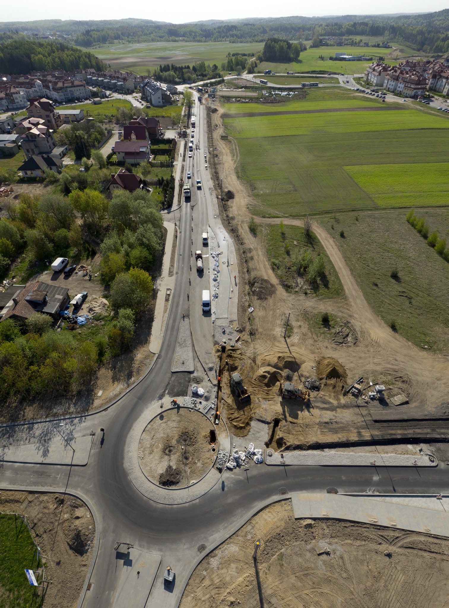 Widok z drona na budowę ronda i nowej drogi w obszarze zabudowanym. W tle widoczne domy, pola i las.