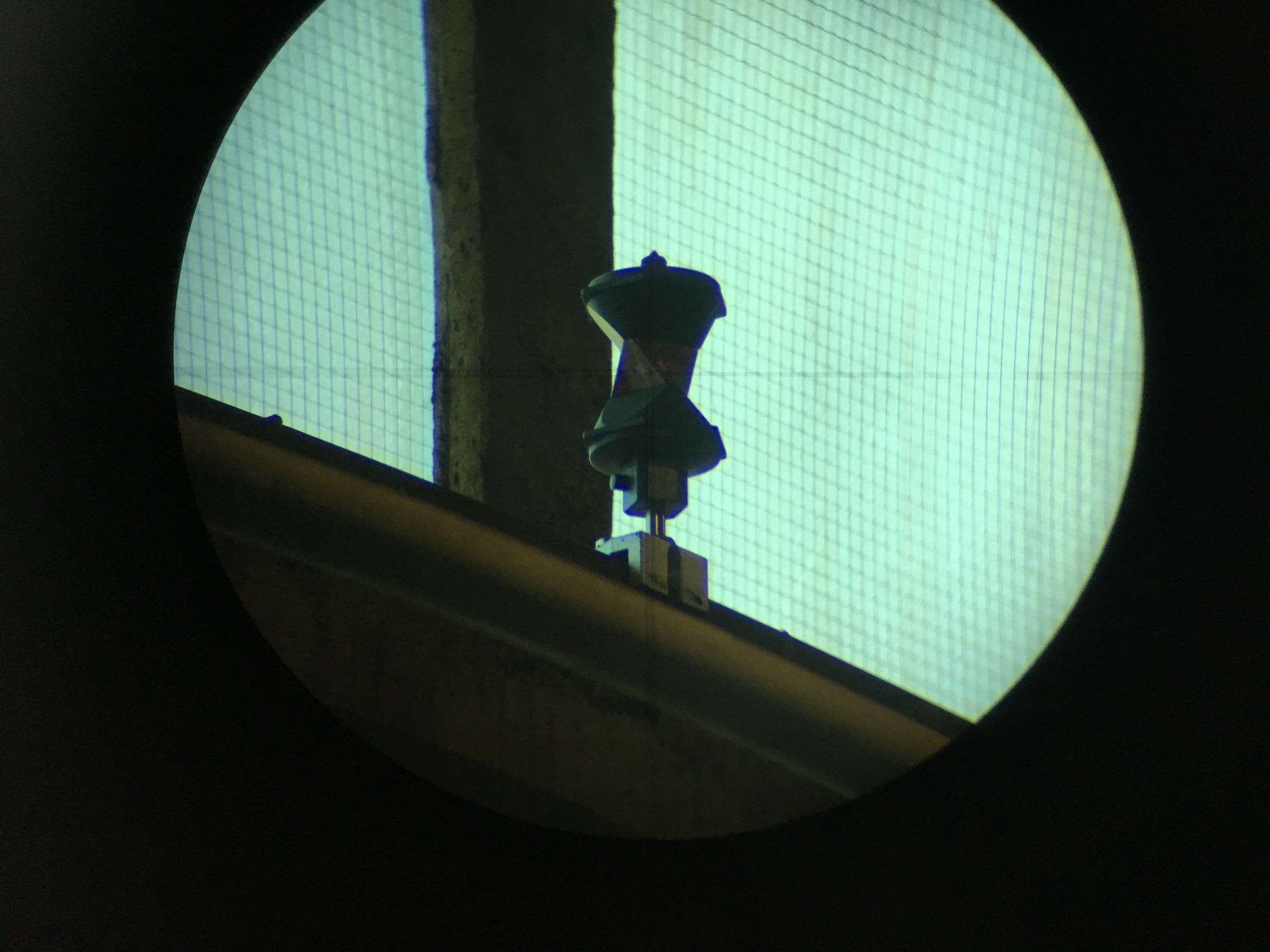 Widok przez teodolit na cel geodezyjny na budynku z siatką ochronną w tle.