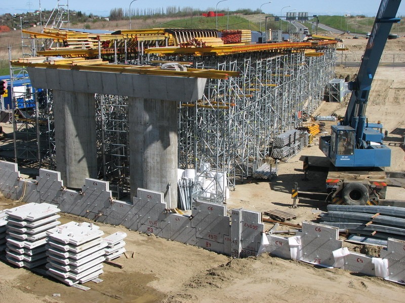 Budowa wiaduktu: betonowe filary w trakcie wznoszenia, rusztowania, dźwig i ułożone płyty betonowe z oznaczeniami na placu budowy.