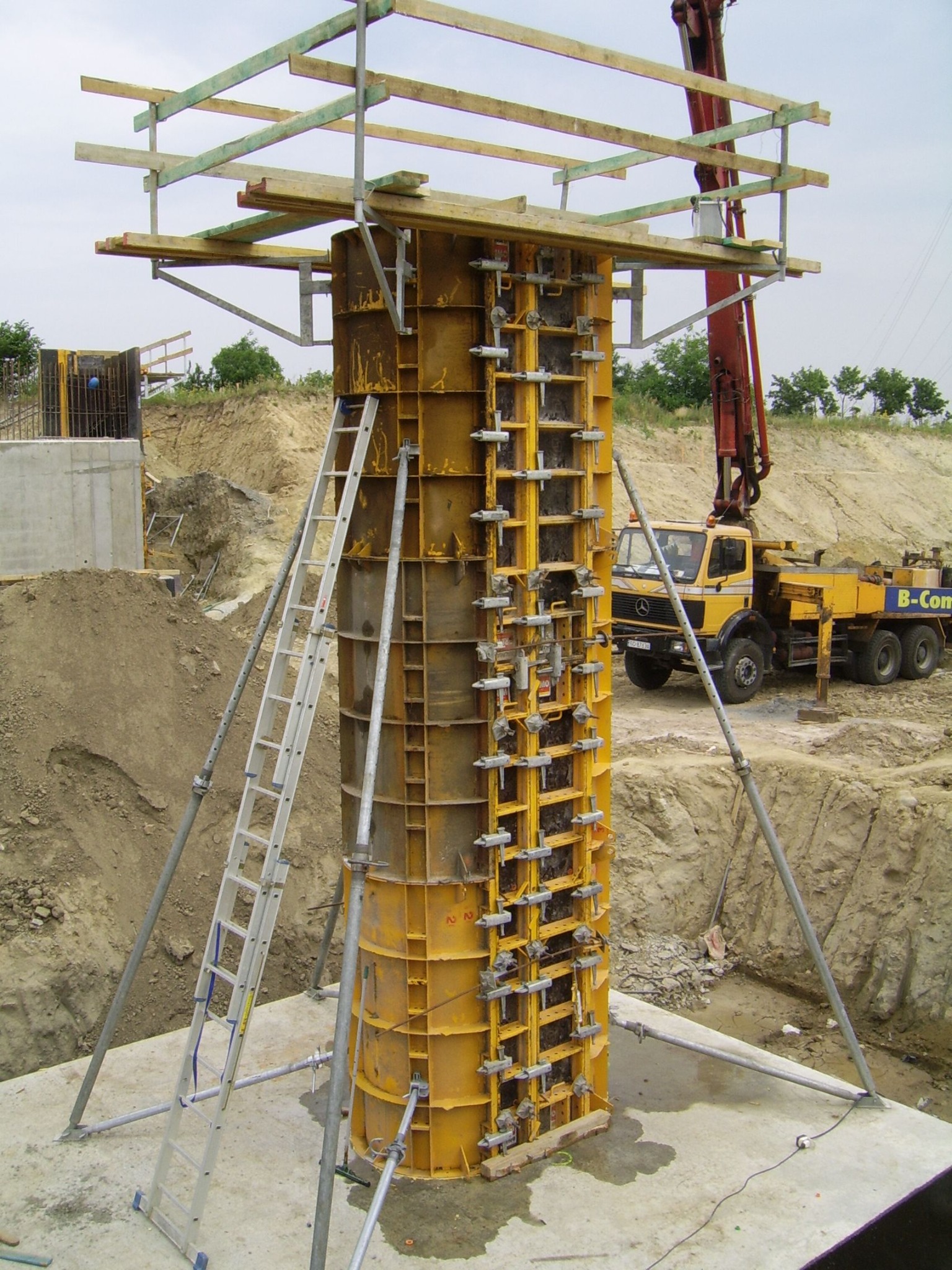 Żółty szalunek kolumny wzmocniony metalowymi wspornikami i drabiną na placu budowy, w tle pompa do betonu na podwoziu Mercedesa