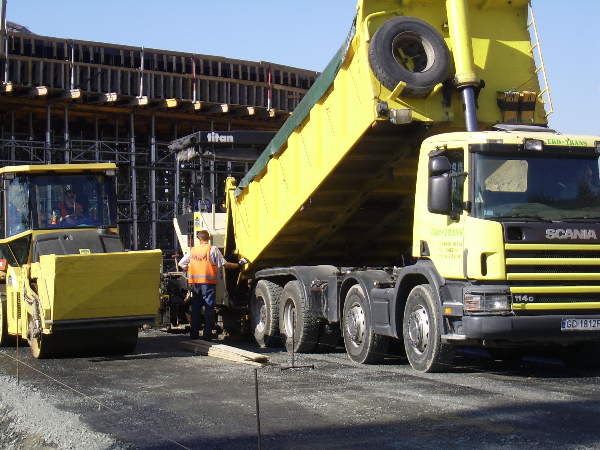 Żółta wywrotka Scania EKO-TRANS w trakcie rozładunku materiału na budowie, obok walec drogowy i pracownik w pomarańczowej kamizelce odblaskowej, w tle szalunki konstrukcji betonowej.