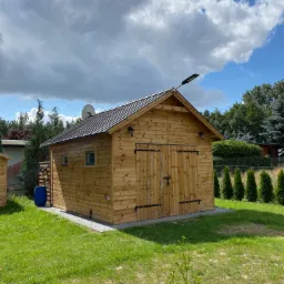 Drewniana konstrukcja z dwuskrzydłowymi drzwiami i metalowym dachem, oświetlona lampą solarną, z satelitą na dachu, stojąca na trawniku w słoneczny dzień.