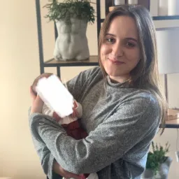 Uśmiechnięta kobieta w szarym swetrze trzyma na rękach niemowlę z zamazaną twarzą, w tle półki z dekoracjami i roślinami w doniczkach w kształcie popiersia.