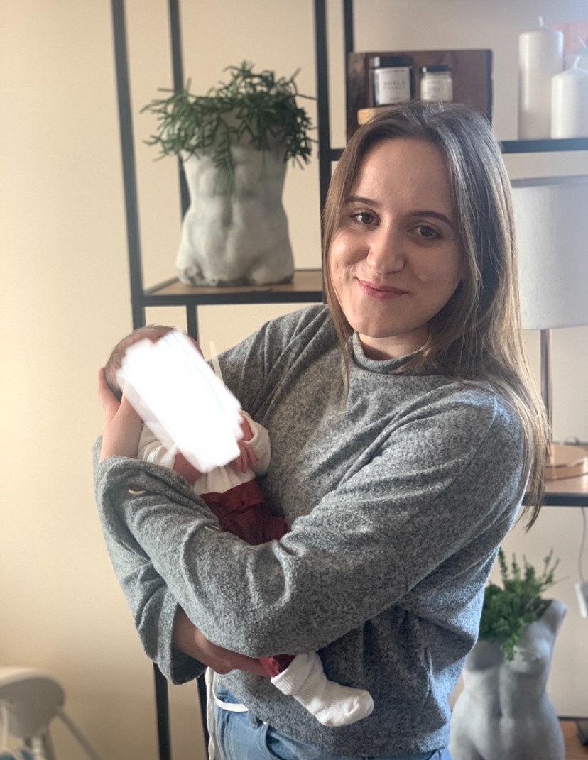 Uśmiechnięta kobieta w szarym swetrze trzyma na rękach niemowlę z zamazaną twarzą, w tle półki z dekoracjami i roślinami w doniczkach w kształcie popiersia.