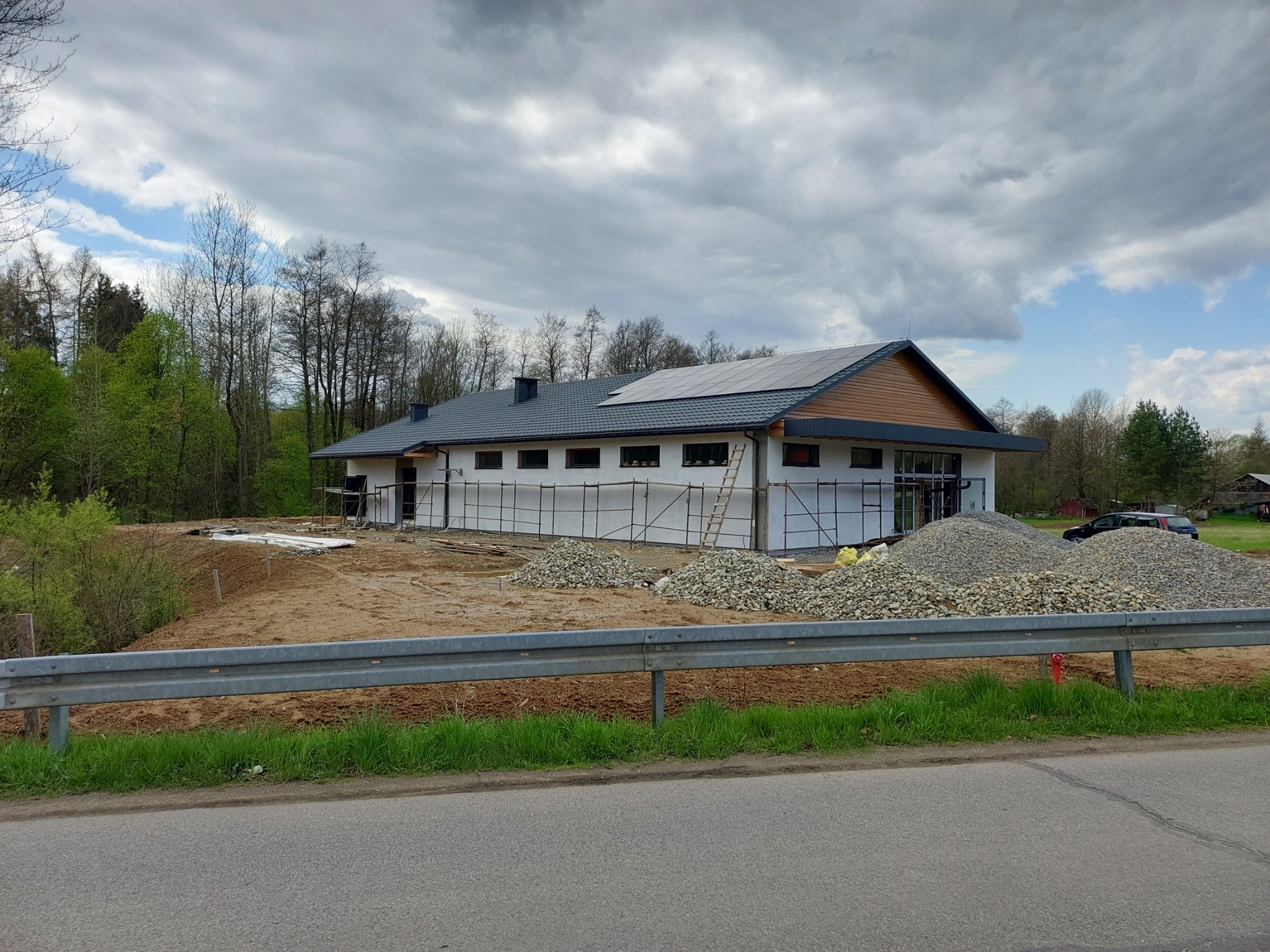 Budynek w trakcie budowy z panelami słonecznymi na dachu, otoczony rusztowaniami i stertami żwiru, na tle zachmurzonego nieba.