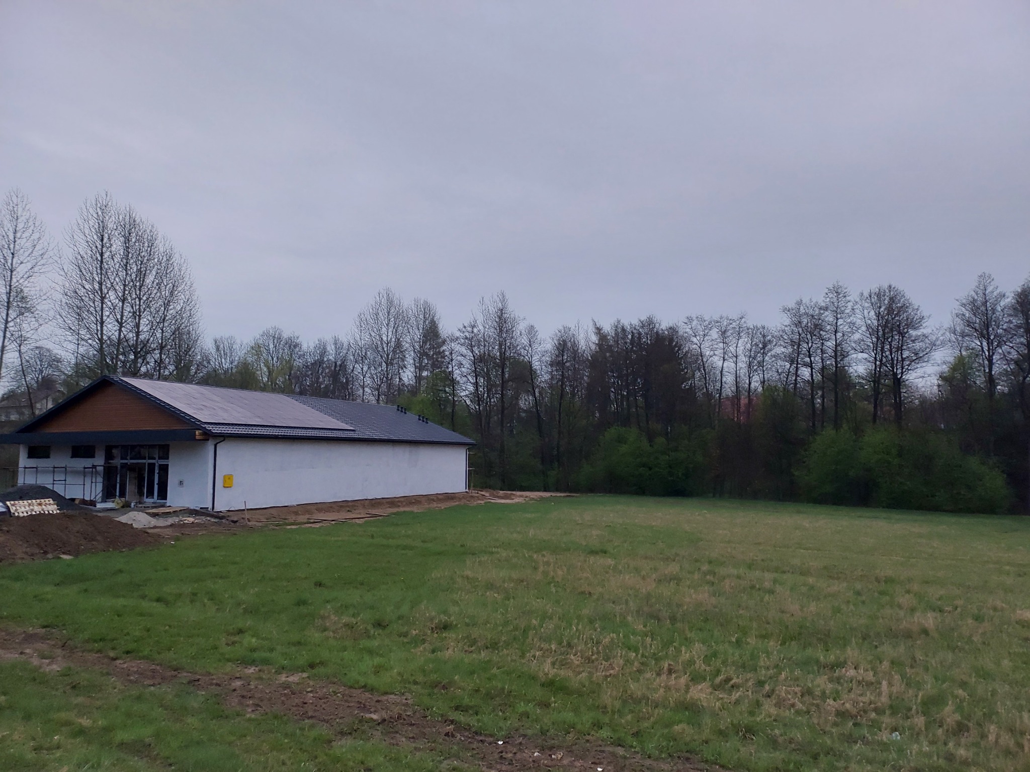 Biały budynek z brązowym dachem i panelami fotowoltaicznymi w pochmurny dzień, widok z trawnika.