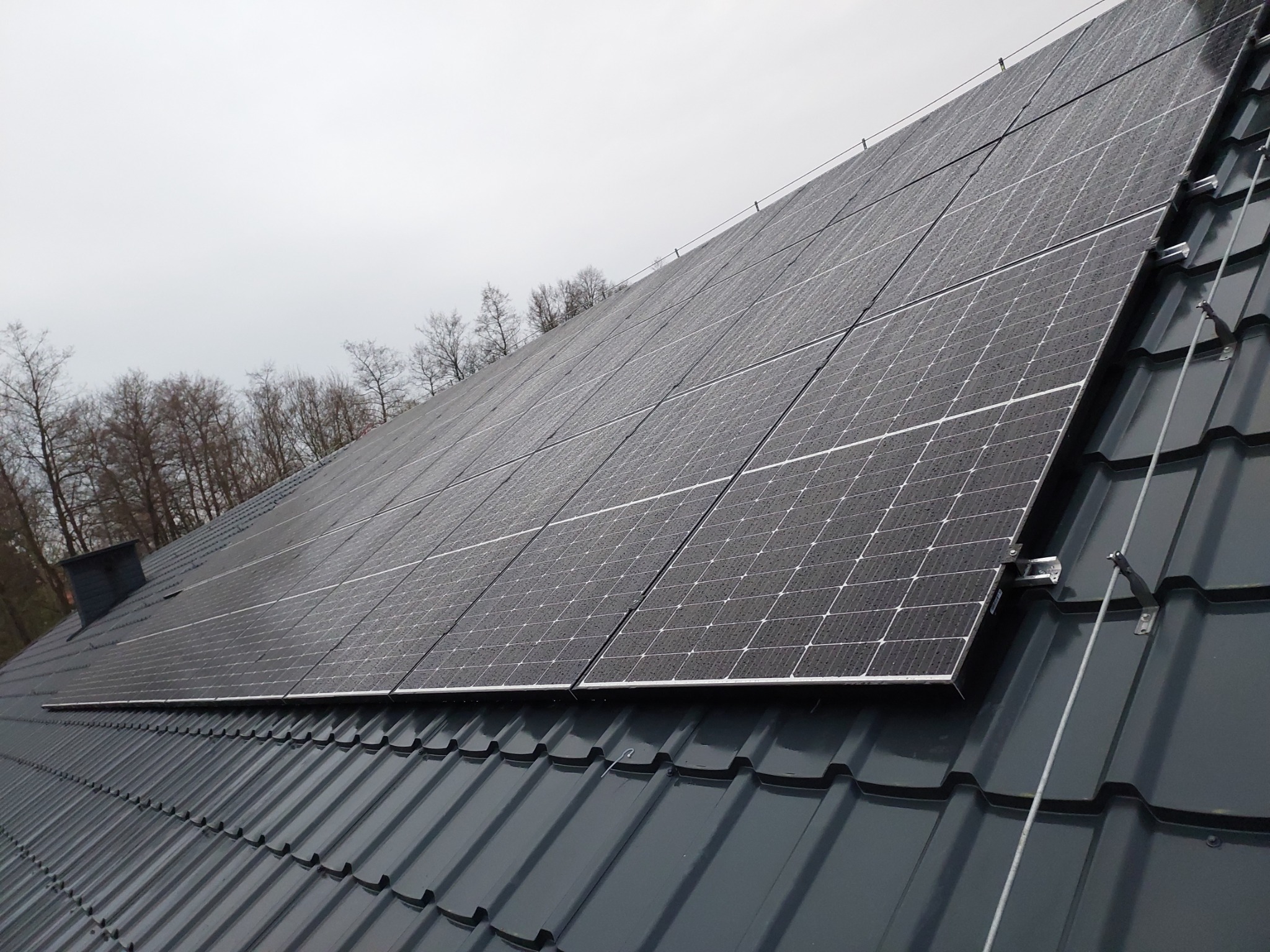 Ciemne panele słoneczne na grafitowym dachu z blachodachówki w pochmurny dzień, widoczne krople deszczu na panelach.