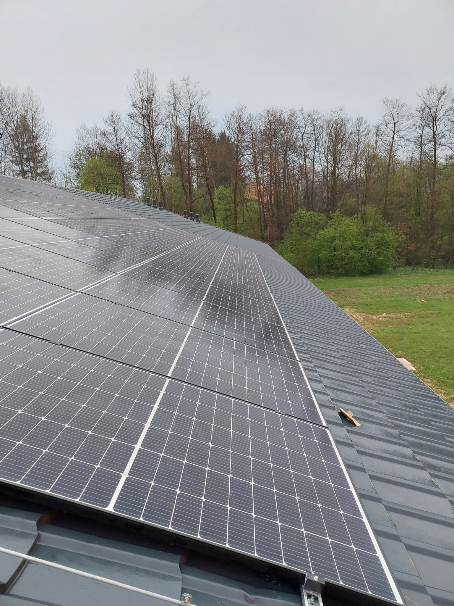 Panel słoneczny na dachu z szarej dachówki, widok na drzewa w tle i fragment trawnika.