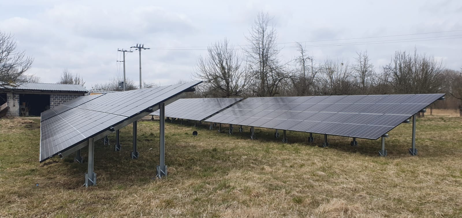 Instalacja paneli słonecznych na metalowych konstrukcjach na zewnątrz, widok na pobliski budynek i słupy energetyczne w tle.