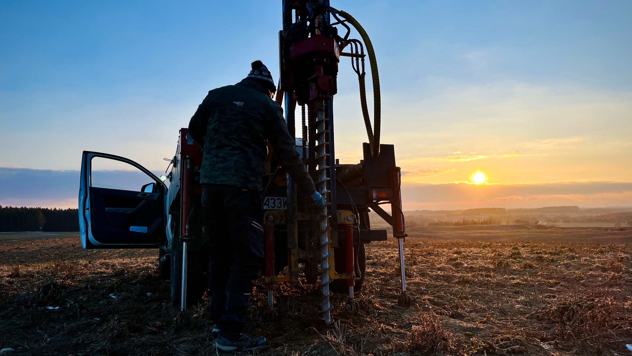 Prace geologiczne w terenie: mężczyzna obsługuje wiertnicę, w tle otwarte drzwi samochodu i zachodzące słońce nad polami. Profesjonalny sprzęt w akcji.
