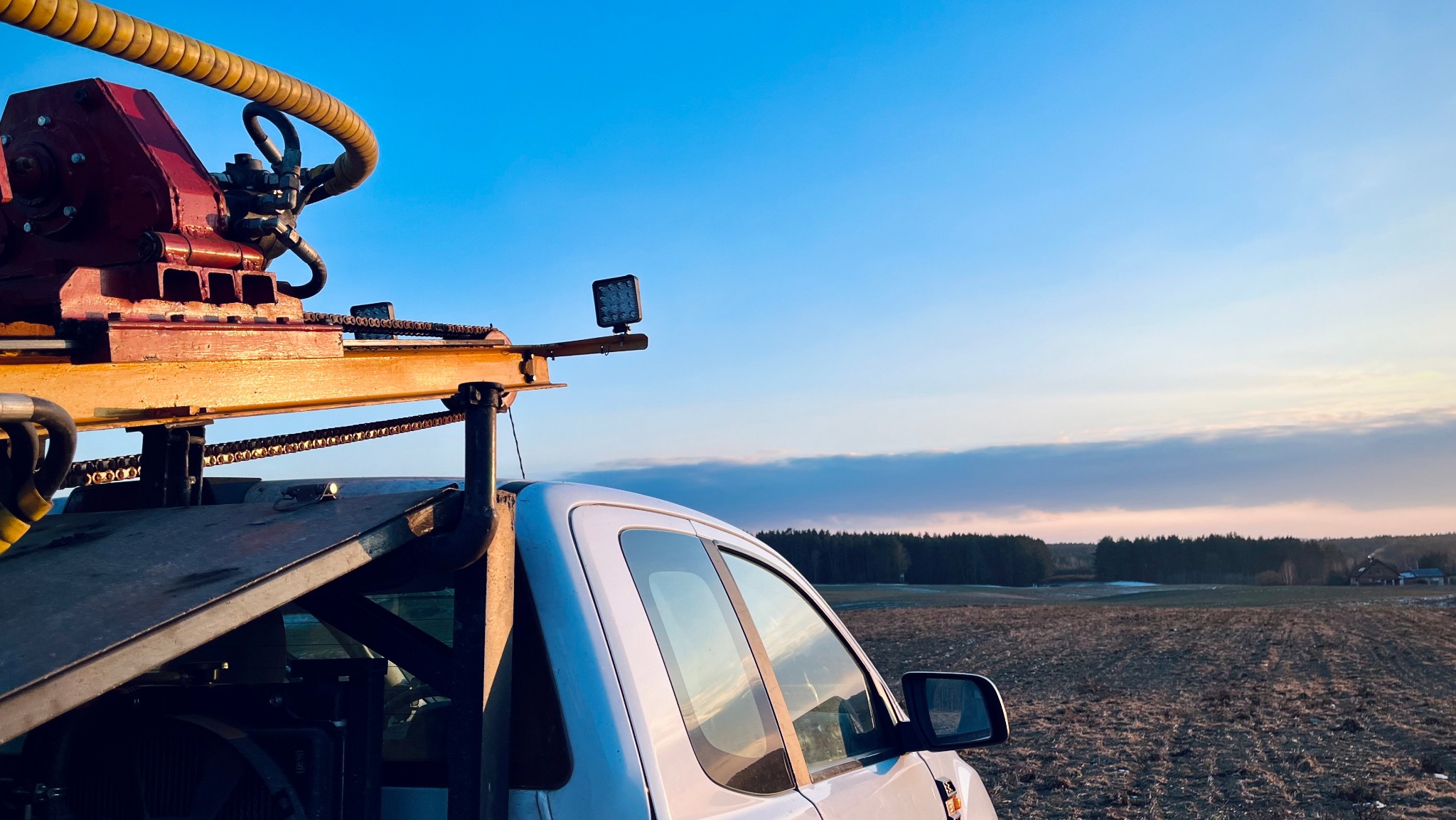 Wiertnica zamontowana na samochodzie terenowym na tle pola i lasu o zmierzchu. Specjalistyczny sprzęt do wierceń geologicznych w terenie.