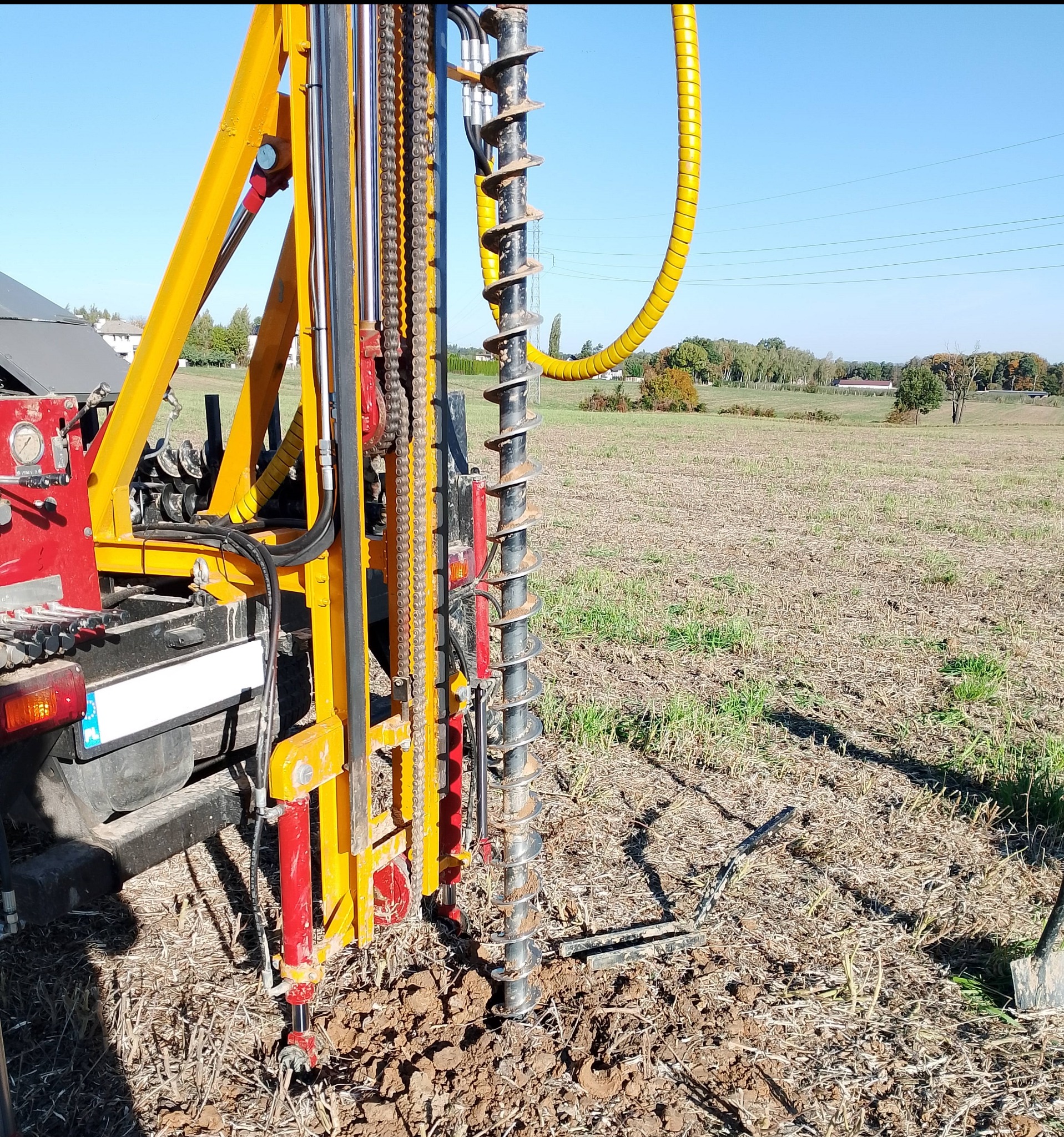 Żółto-czerwona wiertnica geologiczna w trakcie wiercenia otworu w ziemi na tle pola i błękitnego nieba. Widoczne narzędzia i elementy maszyny.