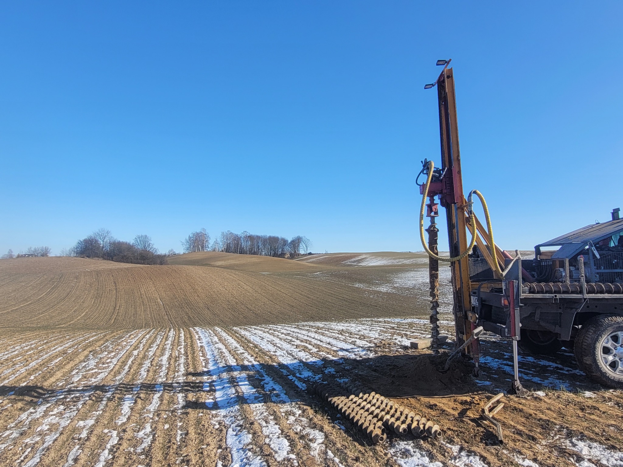 Wiertnica geologiczna w trakcie pracy na polu z resztkami śniegu, błękitne niebo w tle. Widoczne wiertła i fragment ciężarówki. Badanie gruntu w zimowej scenerii.
