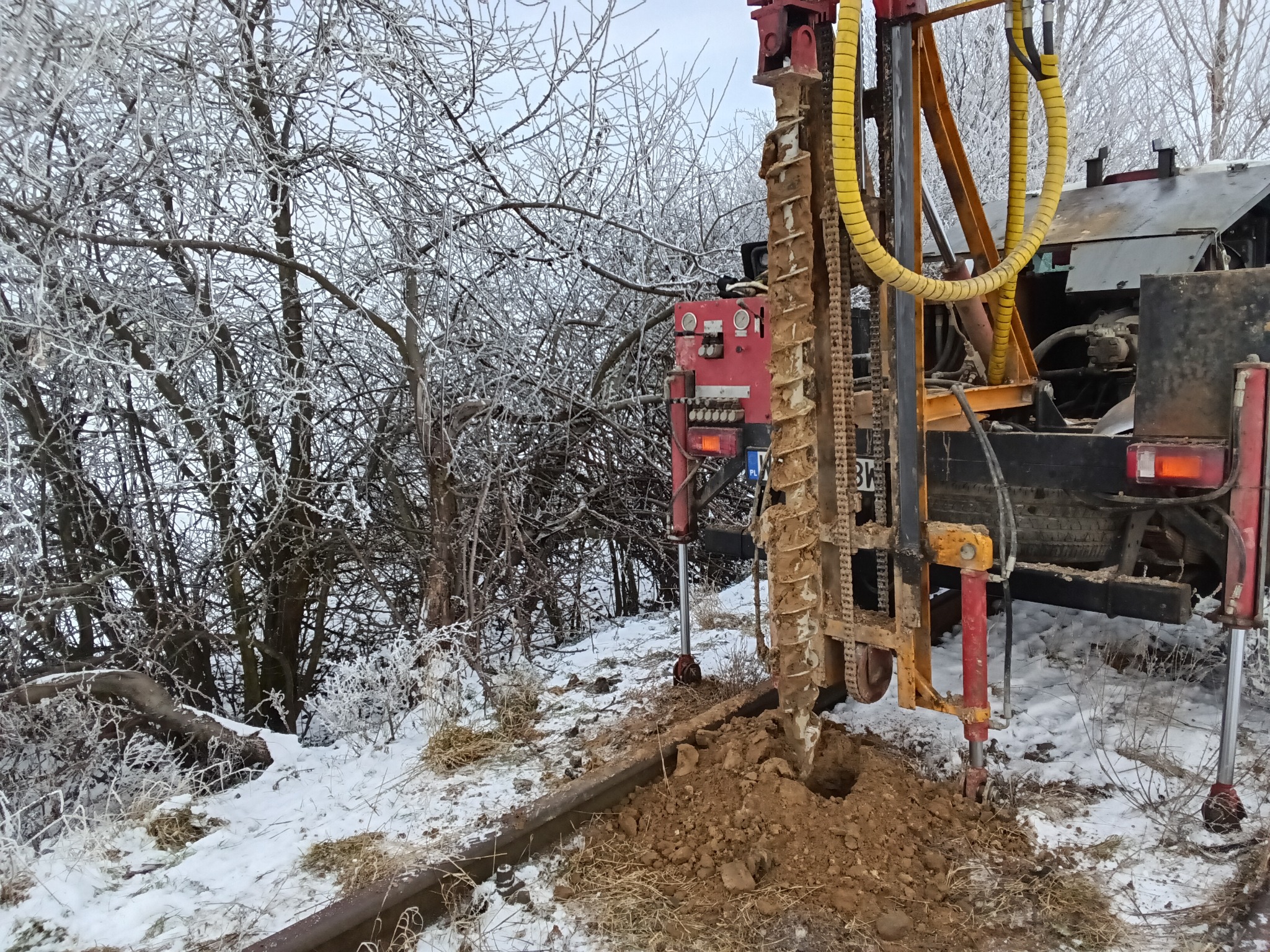 Wiertnica w zimowej scenerii, wiercąca otwór w ziemi. Śnieg i oszronione gałęzie drzew w tle. Widoczny fragment wiertła i mechanizm wiertniczy.