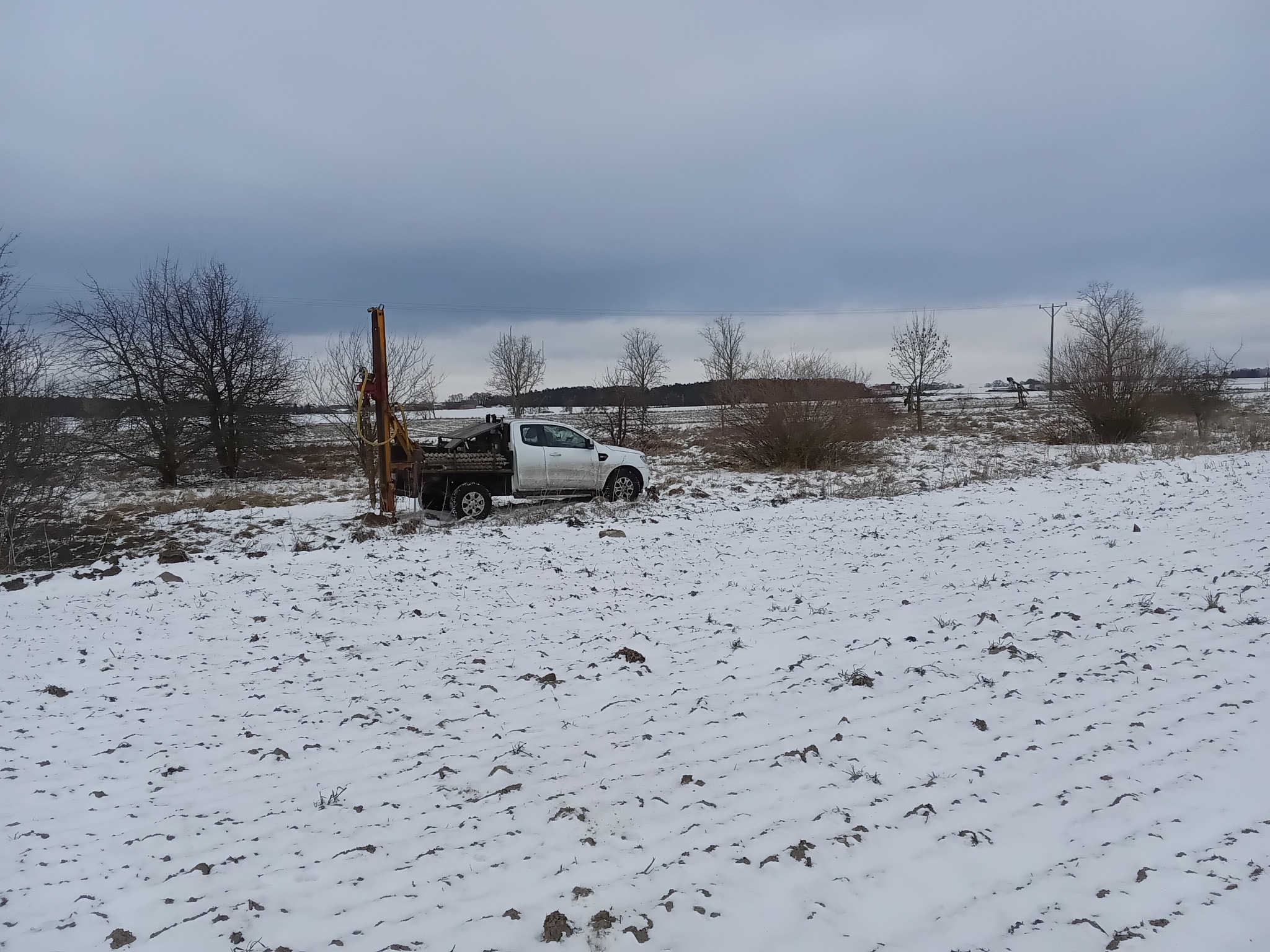 Prace geologiczne w zimowej scenerii Zielonki. Biały samochód terenowy z wiertnicą na zasypanym śniegiem polu. Pochmurne niebo.