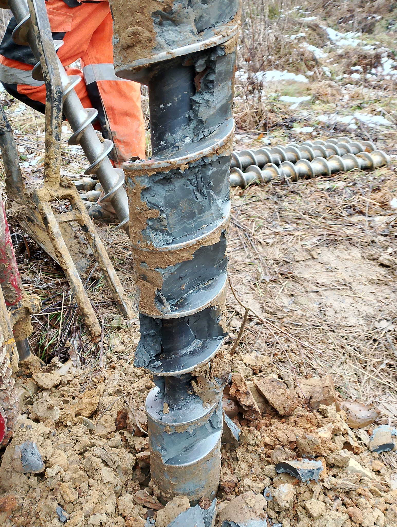 Wiertło świdrowe w ziemi, pokryte błotem, używane podczas wierceń geologicznych. W tle widoczne kolejne wiertła i fragment pomarańczowego kombinezonu.