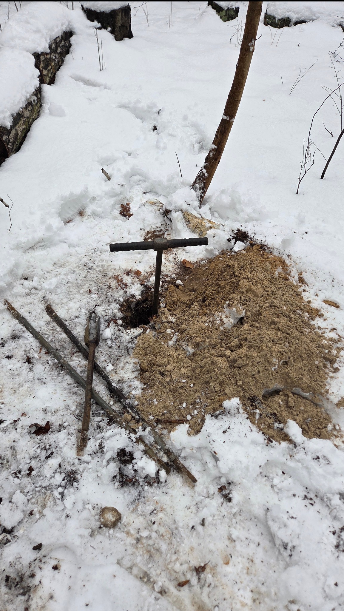 Wykonywanie odwiertu w zasypanym śniegiem terenie. Wokół leżą narzędzia, a obok otworu usypana jest ziemia. Widoczne ślady pracy w zimowych warunkach.