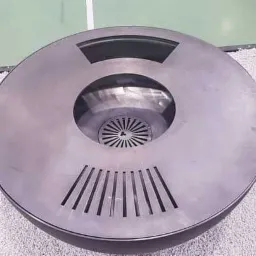 Okrągła, metalowa fontanna z centralnym otworem i promienistym wzorem na dnie, otoczona białym żwirem i ujęta w drewnianą ramę, na tle zielonej podłogi.