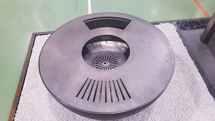 Okrągła, metalowa fontanna z centralnym otworem i promienistym wzorem na dnie, otoczona białym żwirem i ujęta w drewnianą ramę, na tle zielonej podłogi.