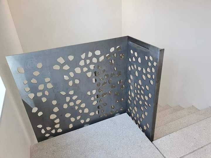 Metalowa barierka schodowa z dekoracyjnymi, nieregularnymi otworami, zamontowana na betonowych stopniach w jasnym wnętrzu.