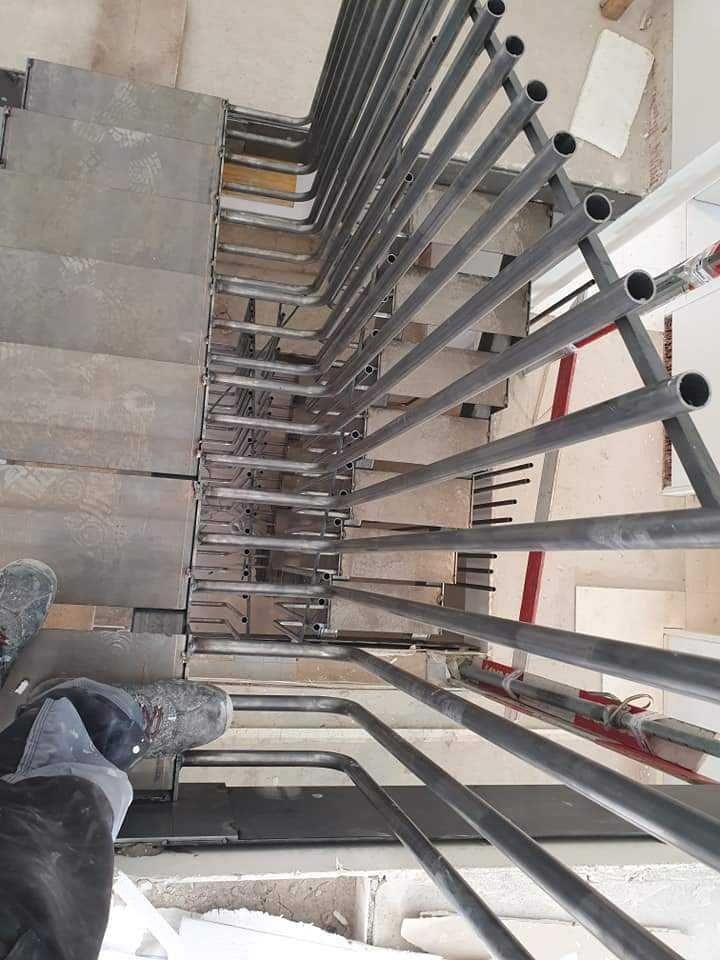 Widok z góry na konstrukcję stalową schodów z surowymi, przyspawanymi poręczami z rur, częściowo zamontowaną na betonowych stopniach. Widoczne buty montera w dolnym rogu kadru.