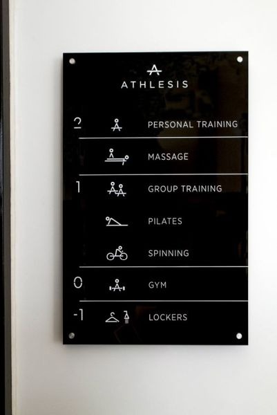 Czarna tabliczka informacyjna z białymi piktogramami i napisami, prezentująca ofertę usług klubu fitness: trening personalny, masaż, trening grupowy, pilates, spinning, siłownia, szatnia.