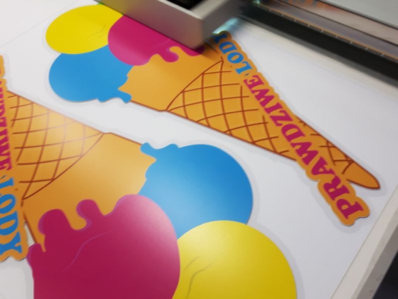 Proces wycinania naklejek w kształcie lodów z napisem 'Prawdziwe lody' w jaskrawych kolorach, widoczna głowica tnąca maszyny.