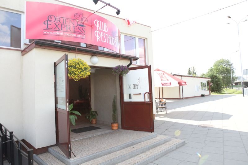 Wejście do restauracji Orient Express z otwartymi drzwiami, wiszącymi kwiatami i reklamowym banerem nad wejściem.