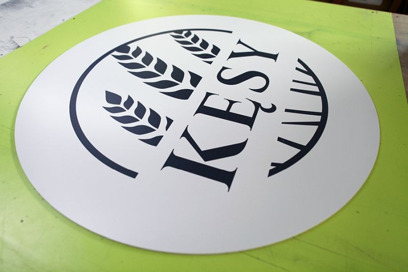 Okrągła naklejka z logo 'KESY' i motywem trzech kłosów zboża, umieszczona na zielonym podłożu.