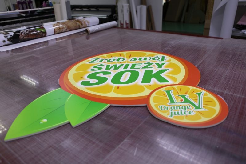 Dwa wycięte z tworzywa sztucznego, okrągłe szyldy reklamowe z grafiką i napisem 'Zrób swój ŚWIEŻY SOK' oraz logo 'Orange juice', leżące na stole w drukarni, w tle widoczne rolki z wydrukami.