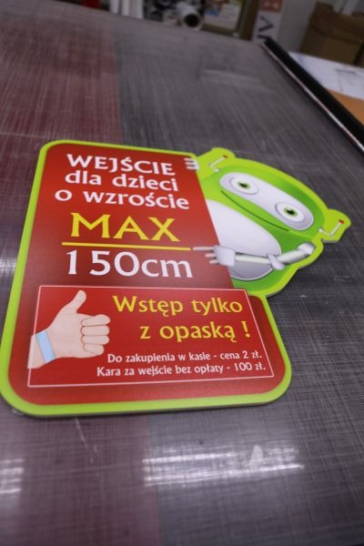 Tabliczka informacyjna z ograniczeniem wzrostu dla dzieci do 150 cm, z zielonym obrysem i elementem graficznym przypominającym robota, umieszczona na szarej powierzchni.
