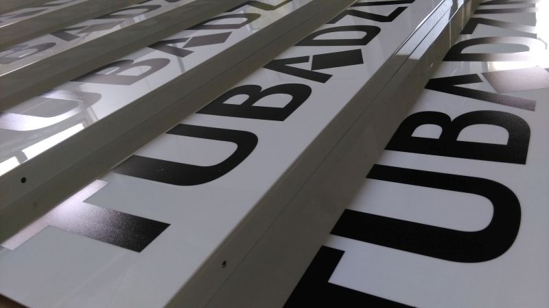 Rząd białych tablic z czarnymi literami 'TUBADZ...' w perspektywie, ułożonych jedna za drugą, gotowych do montażu.