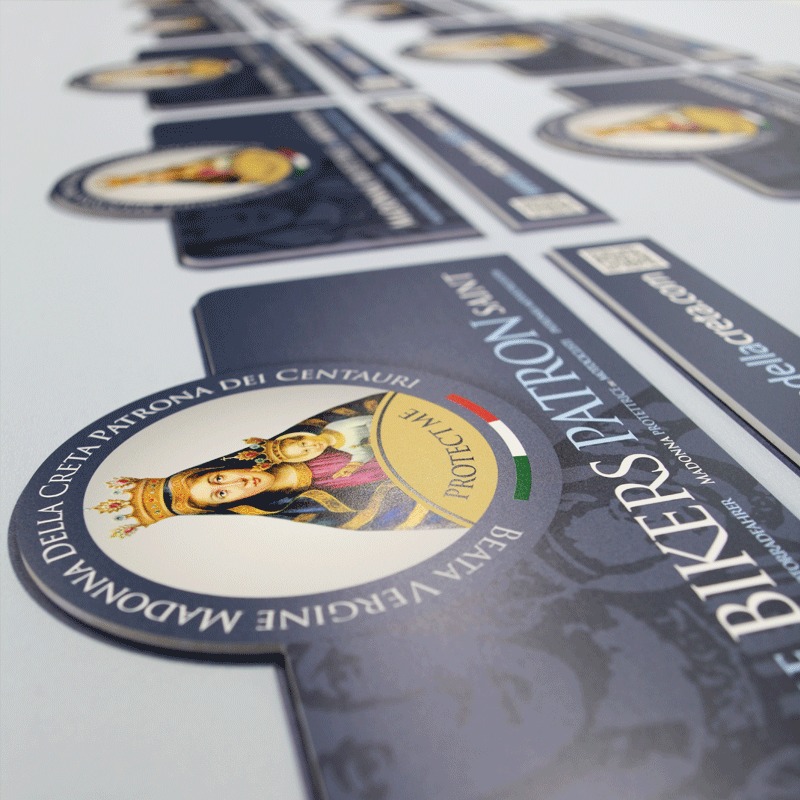 Arkusze z wyciętymi naklejkami i etykietami z motywem religijnym i logo 'Bikers Patron', widoczne z bliska pod kątem, na białym tle.