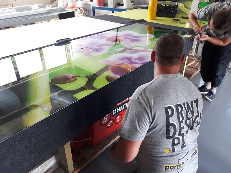 Przycinanie nożykiem grafiki z motywem kwiatowym na szkle, widoczna osoba w koszulce z napisem 'PRINT DESIGN PL' oraz odbicie okna w szkle.