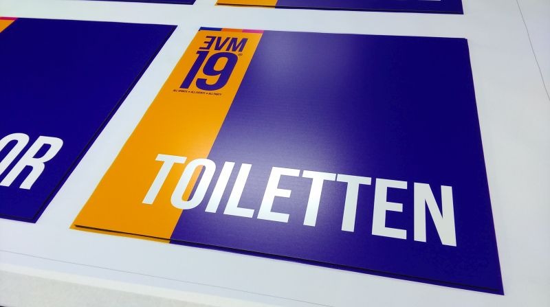 Niebiesko-żółte tablice informacyjne z błyszczącym napisem 'TOILETTEN' i logo '3VM 19' w górnym rogu, ułożone na białym podłożu.