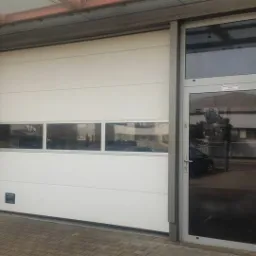 Biała segmentowa brama garażowa z oknami w górnej części, zamontowana obok przeszklonych drzwi wejściowych z aluminiową ramą, widok z zewnątrz budynku.