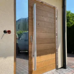 Nowoczesne drzwi zewnętrzne z drewnianymi panelami i lustrzanym bocznym elementem, jeszcze z zabezpieczeniem transportowym, widoczne przewody elektryczne wystające ze ściany.