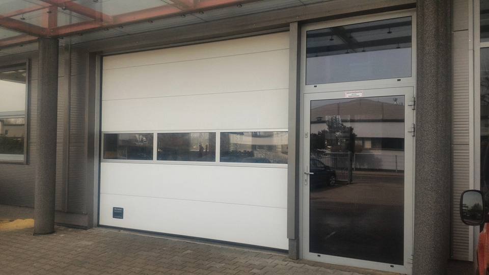 Biała segmentowa brama garażowa z oknami w górnej części, zamontowana obok przeszklonych drzwi wejściowych z aluminiową ramą, widok z zewnątrz budynku.