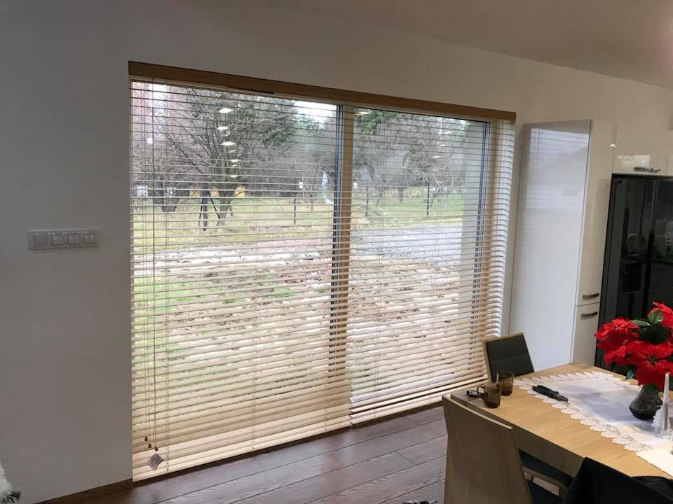 Drewniane żaluzje poziome w jasnym odcieniu, zamontowane w dużym oknie z widokiem na ogród w pochmurny dzień, widoczne elementy wyposażenia wnętrza: stół z dekoracją, krzesła, biała szafka.