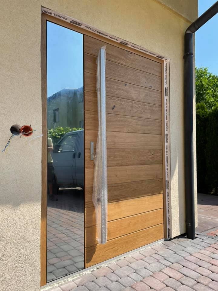 Nowoczesne drzwi zewnętrzne z drewnianymi panelami i lustrzanym bocznym elementem, jeszcze z zabezpieczeniem transportowym, widoczne przewody elektryczne wystające ze ściany.