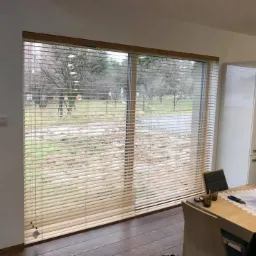 Jasne, drewniane lamele w oknie, widok na ogr&oacute;d z drzewami w tle, część jadalni z drewnianym stołem i czerwonymi kwiatami.