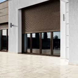 Elewacja budynku z brązowymi roletami zewnętrznymi opuszczonymi na oknach, oświetlenie zewnętrzne w postaci prostokątnych kinkietów, jasna, kamienna posadzka tarasu.