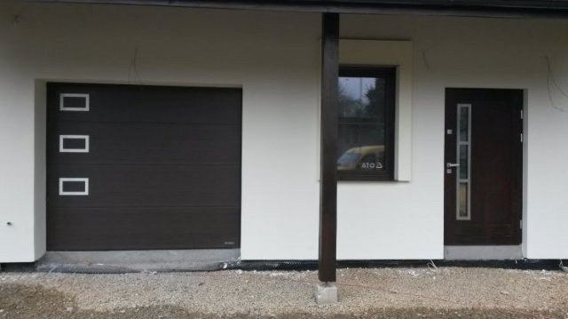 Elewacja domu z brązową segmentową bramą garażową z trzema prostokątnymi okienkami, brązowymi drzwiami wejściowymi z trzema podłużnymi przeszkleniami i oknem z naklejką ATO.