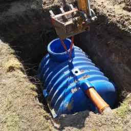 Niebieski, żebrowany zbiornik z rurą drenażową, opuszczany do wykopu za pomocą koparki z czerpakiem zawieszonym na czerwonych pasach.