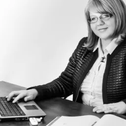 Kobieta w okularach i czarnej kurtce siedzi przy biurku, pisząc na laptopie; obok otwarty segregator; zdjęcie czarno-białe.